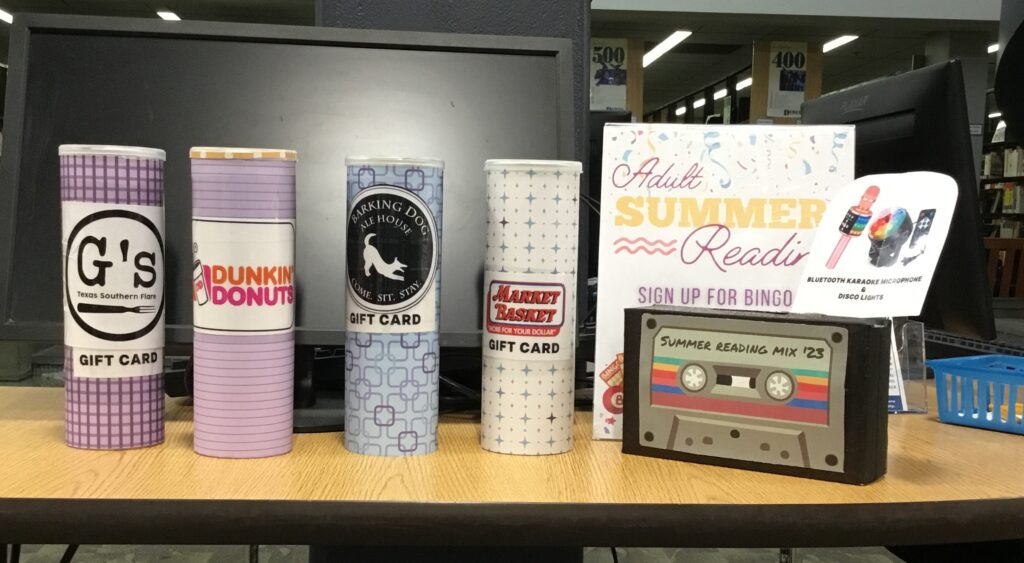 A photo of four tubes resting on a counter, labeled with logos of businesses (G's, Dunkin' Donuts, Barking Dog Ale House, and Market Basket), that serve as buckets to drop tickets in for gift card prizes. To the right of the four tubes is a box with a casette design and the words "Summer Reading Mix '23" on it, and a sign above reading Bluetooth Karaoke Microphone & Disco Lights." This box serves as a bucket to drop tickets in for the Bluetooth karaoke microphone and disco lights prize. Behind this box is a sign that reads "Adult Summer Reading; Sign up for bingo [rest obscured by box]."