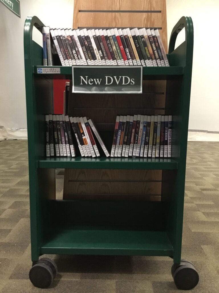 Photo of a green book cart on wheels. The cart has three rows: the top two are filled with DVDs, and the bottom is empty. The cart has a sign that says "New DVDs" attached to it.