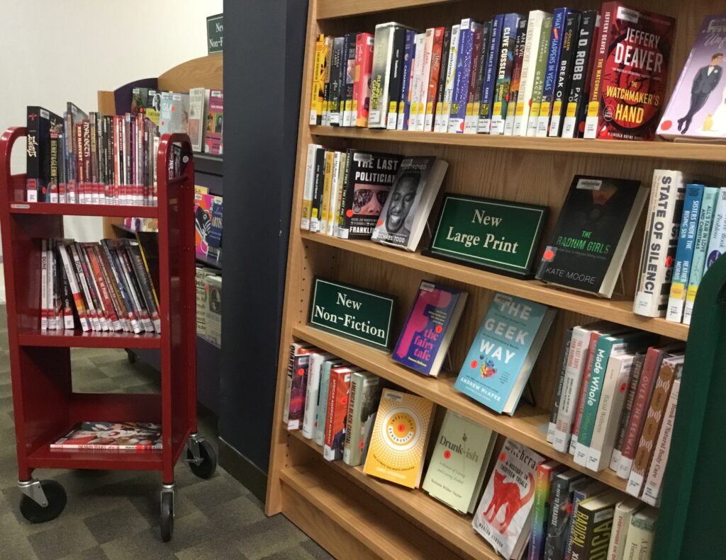 Photo of two bookshelves and a red book cart. The shelf on the left includes a sign, only partially visible, that says "New Non-Fiction." The shelf on the right includes two signs: one that says "New Large Print," and another that says "New Non-Fiction." Both shelves are filled with books. The red cart has three shelves, and holds a variety of graphic novels.