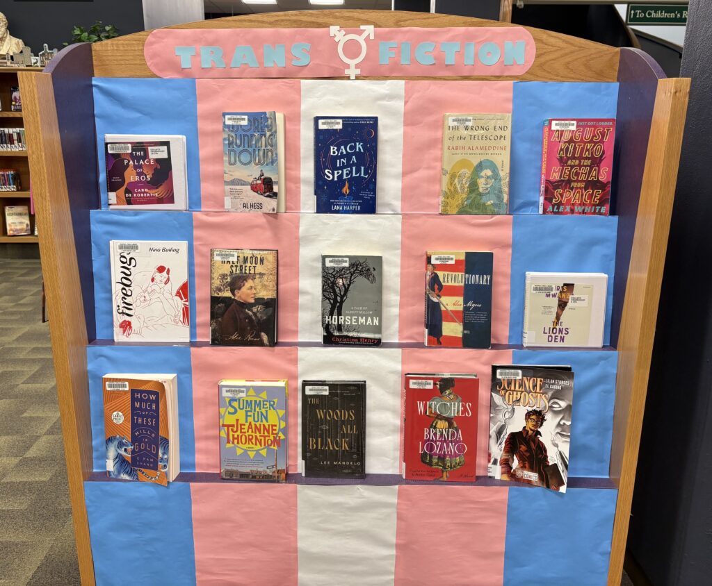 A photo of the adult fiction display for June 2025. It features books propped on a three-level stand, with the colors of the trans flag in the background. At the top it says "Trans Fiction" with a trans symbol in the middle.