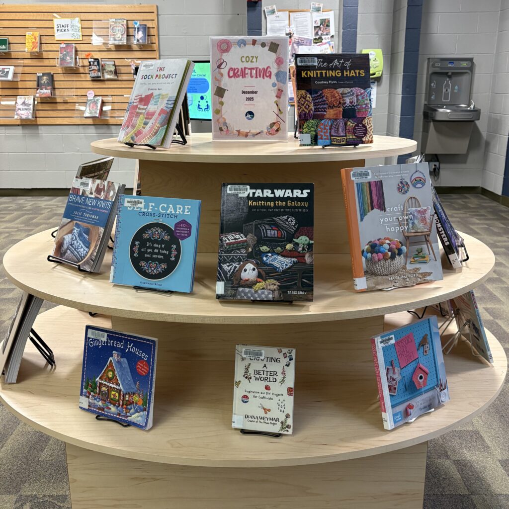 A photo of a set of round shelves filled with crafting-related books on stands. A sign in the middle on the top shelf says "Cozy Crafting December 2025."