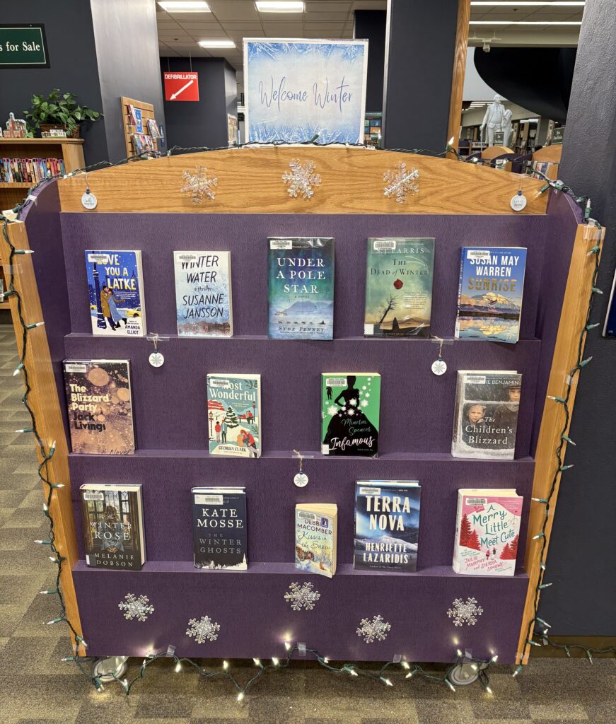 A photo of a set of purple shelves with front-facing books. A sign at the top says "Welcome Winter," and the display includes paper snowflakes and a string of lights around the edge.