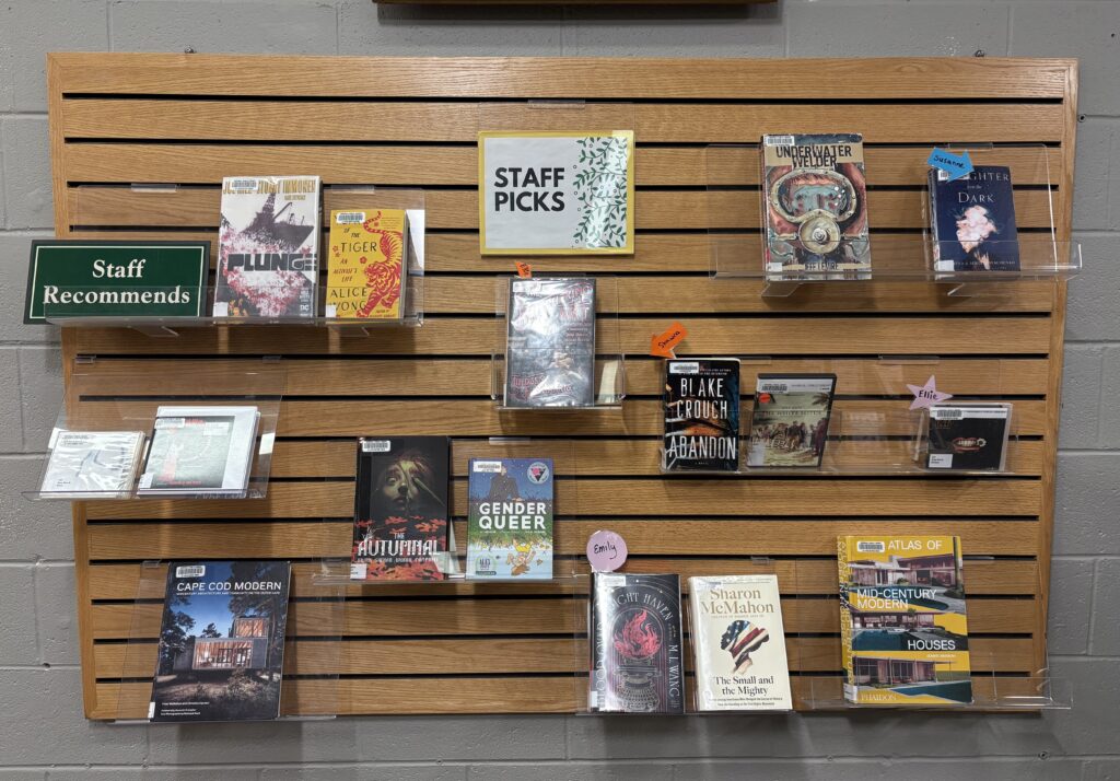 A board on a wall with books on plastic shelves. The board has a sign that says "Staff Picks" at the top in the middle, and another on the top left that says "Staff Recommends."