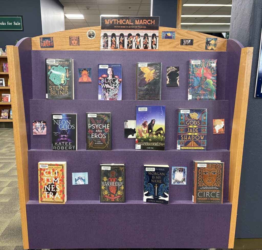 A photo of a set of purple shelves with front-facing books. A sign at the top says "Mythical March," and the display includes paper cutouts with book cover images on them.