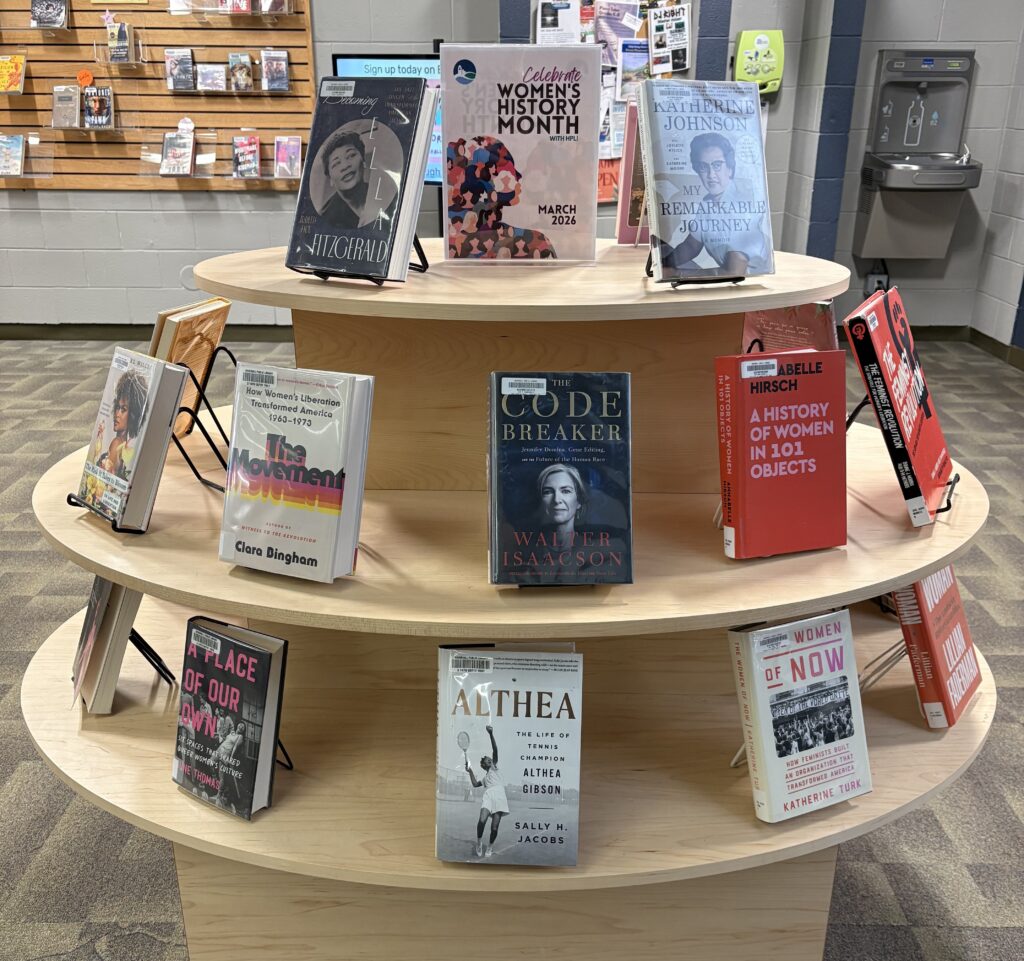A photo of a set of round shelves filled with books related to women's history and biographies of notable women. A sign in the middle on the top shelf says "Celebrate Women's History Month with HPL! March 2026."
