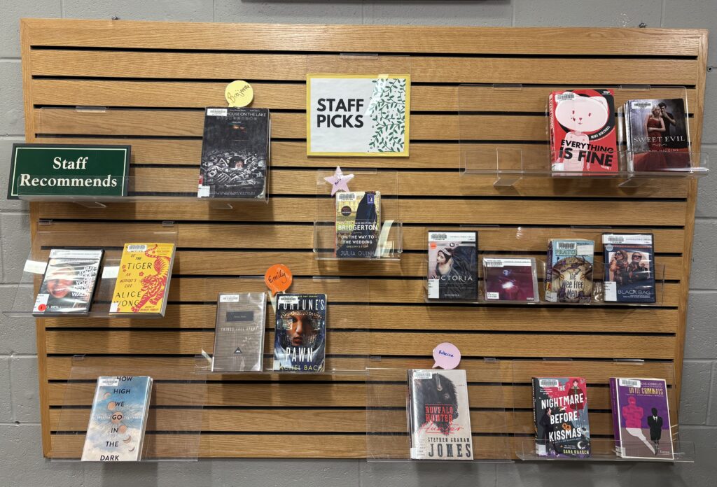 A board on a wall with books on plastic shelves. The board has a sign that says "Staff Picks" at the top in the middle, and another on the top left that says "Staff Recommends."