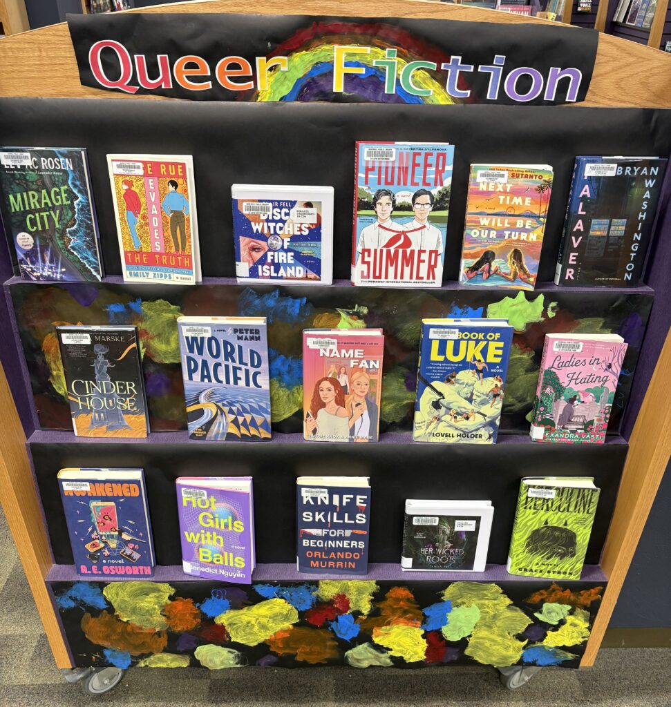 Books on a display shelf with a sign at the top that says "Queer Fiction." The letters are the colors of a rainbow, and a rainbow is painted behind the letters.